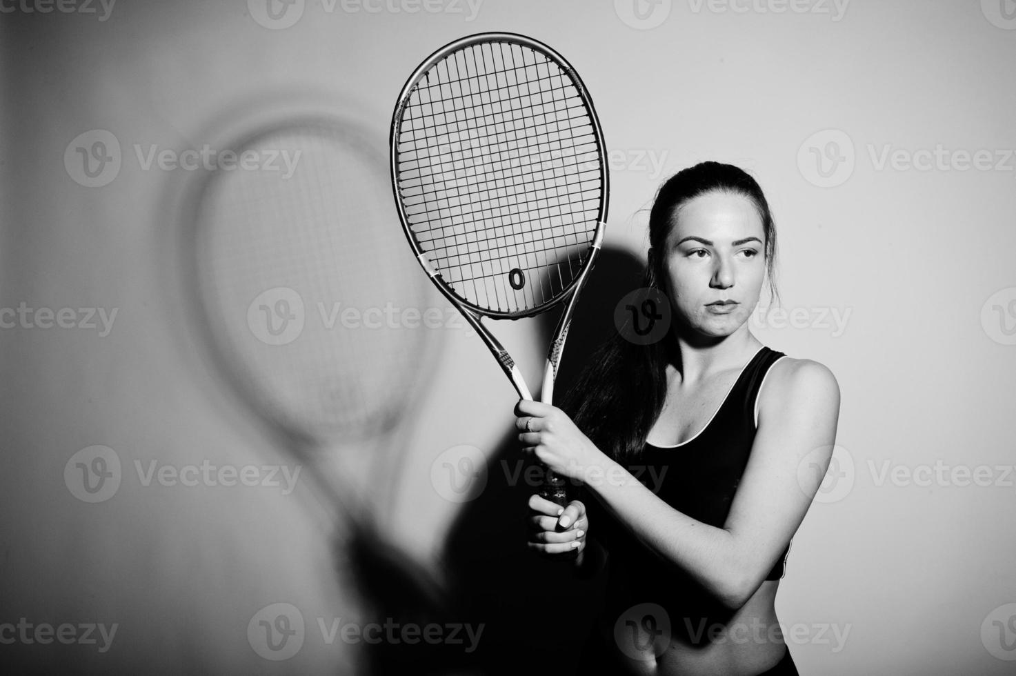 zwart-wit portret van een mooie jonge vrouw in sportkleding die een tennisracket vasthoudt terwijl hij tegen een witte achtergrond staat. foto