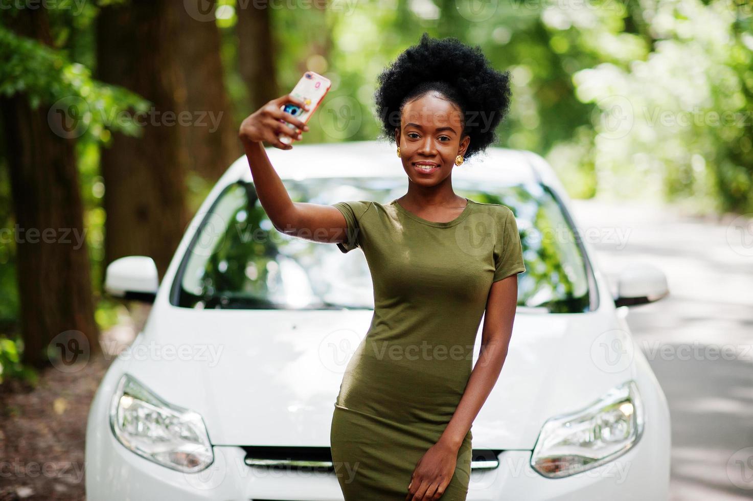 Afro-Amerikaanse vrouw poseerde tegen witte auto in bosweg en keek naar mobiele telefoon. foto