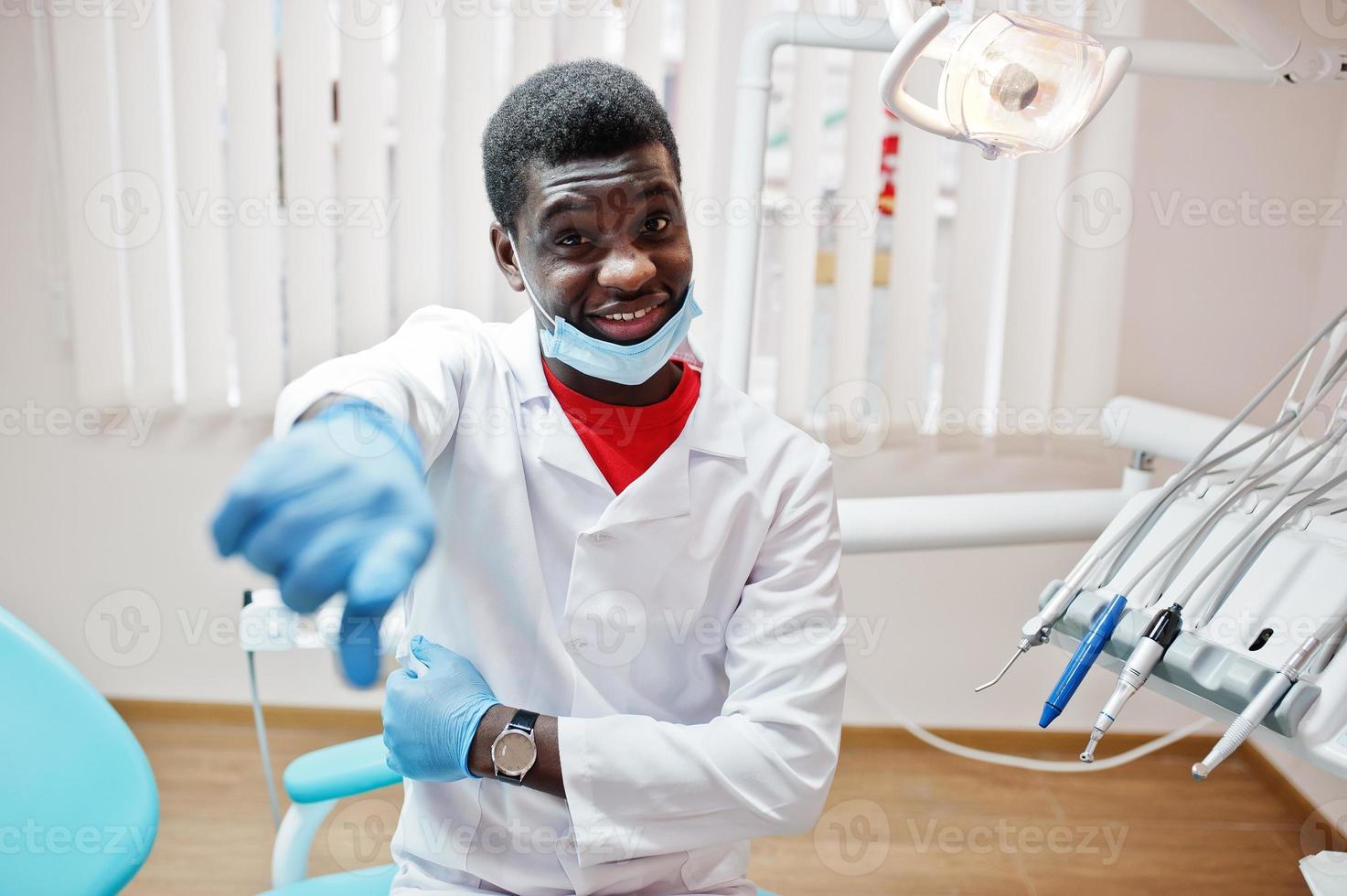 Afro-Amerikaanse mannelijke arts in masker zittend op tandheelkundige kliniek en vinger tonen. foto