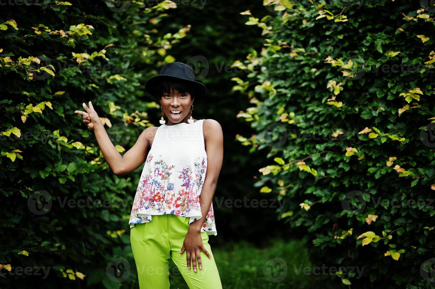 geweldige Afro-Amerikaanse modelvrouw in groene broek en zwarte hoed poseerde met verschillende emoties in het park, toont een twee vingers. foto