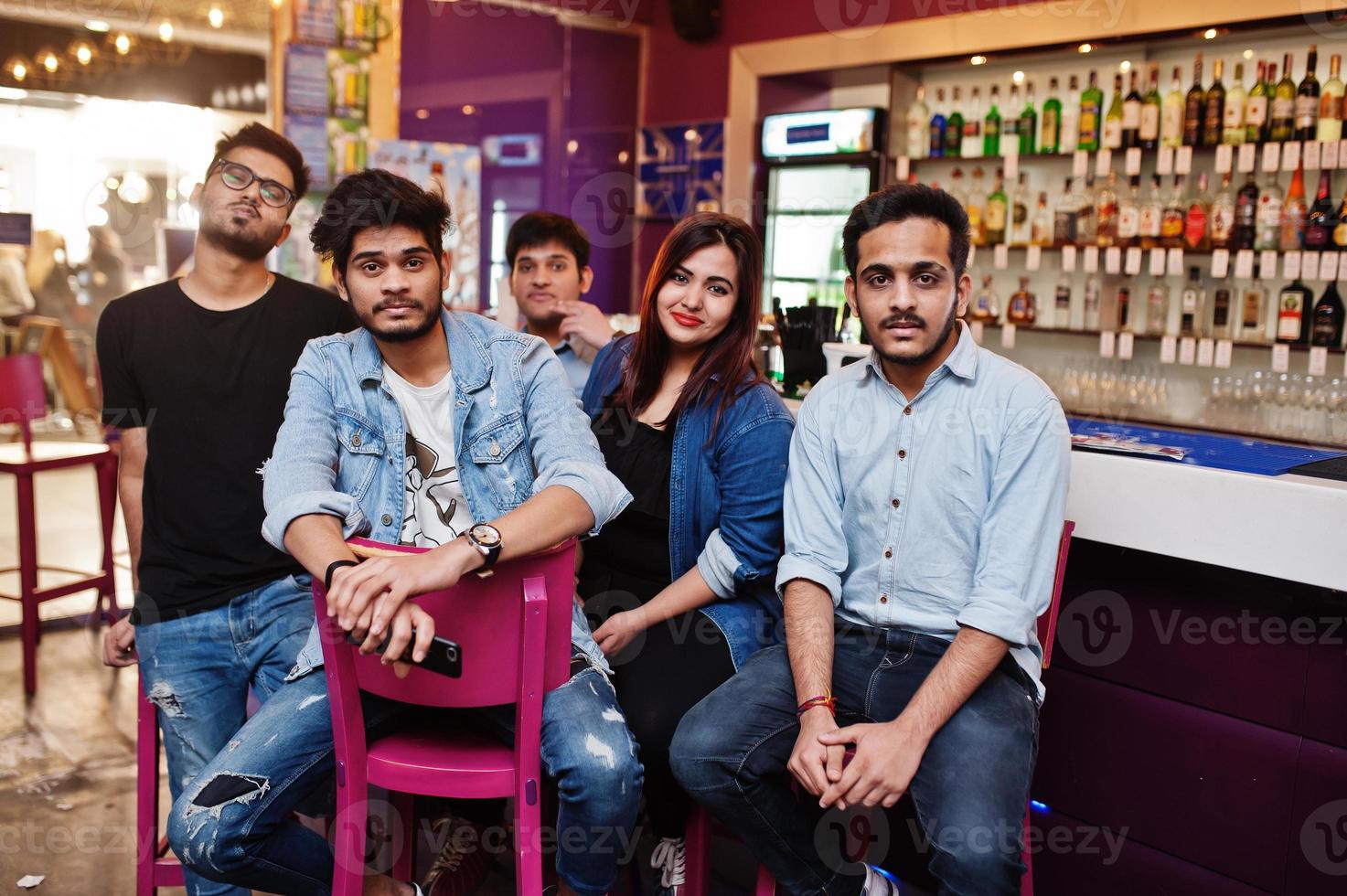 groep stijlvolle aziatische vrienden dragen op jeans zittend op stoelen tegen bar in club. foto