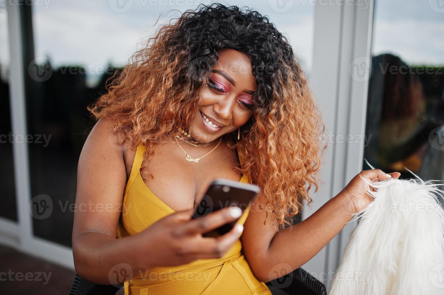 glamour Afro-Amerikaanse vrouw in gele jurk en witte wollen cape zittend op een stoel en kijken op mobiele telefoon. foto