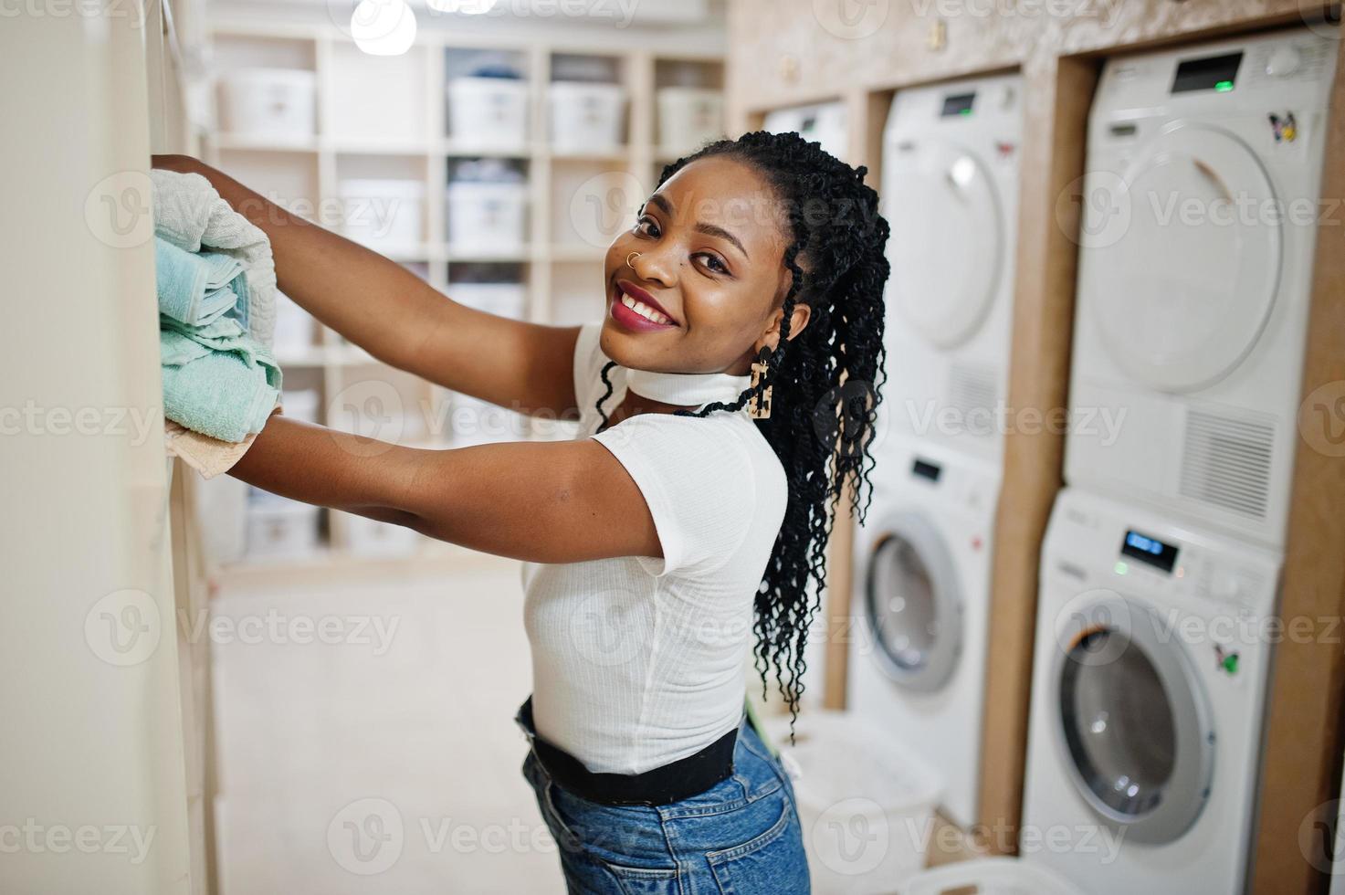 vrolijke Afro-Amerikaanse vrouw met handdoeken in handen in de buurt van wasmachine in de zelfbedieningswasserette. foto