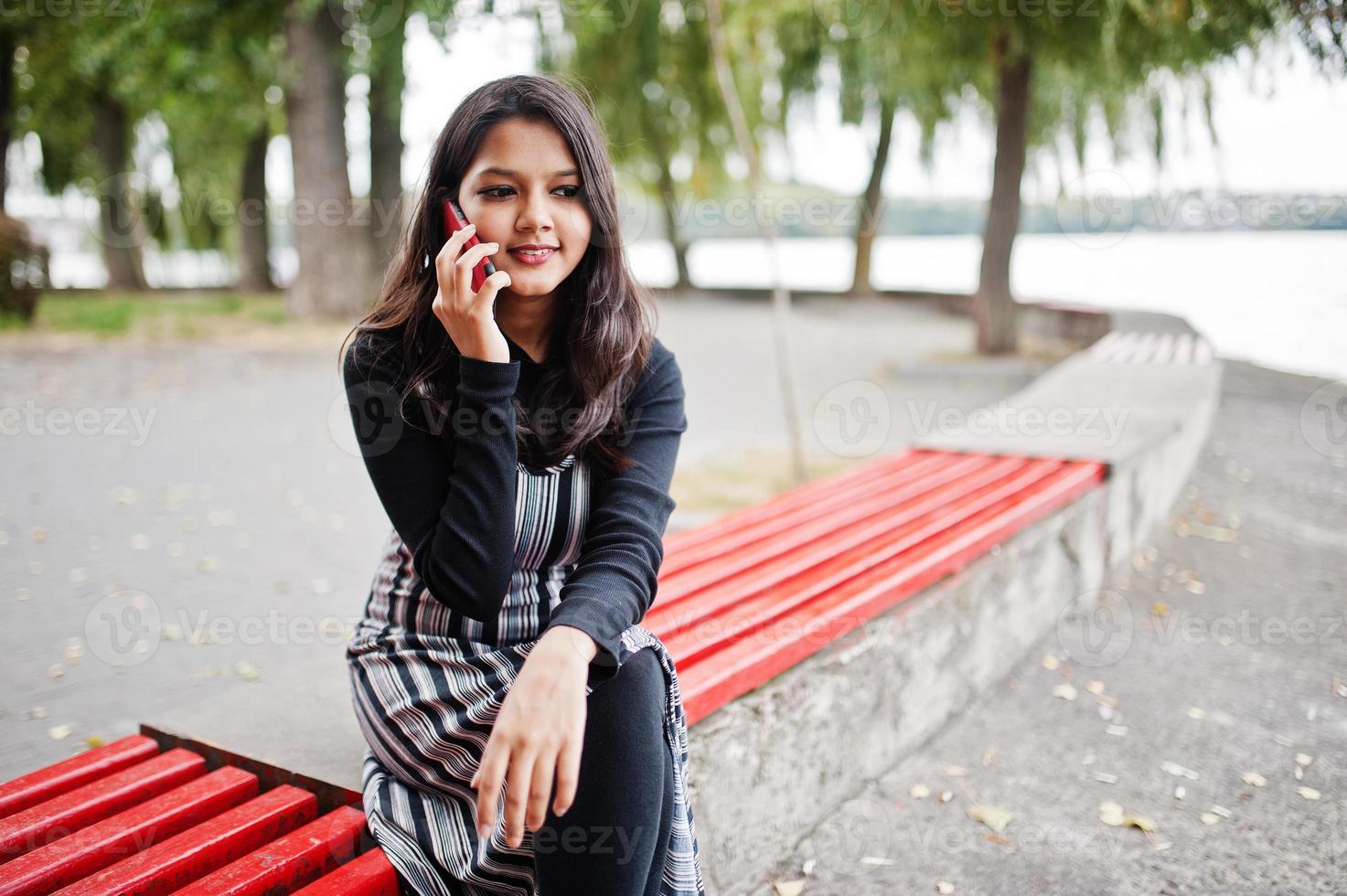 portret van jonge mooie Indiase of Zuid-Aziatische tienermeisje in jurk zittend op de bank met mobiele telefoon. foto