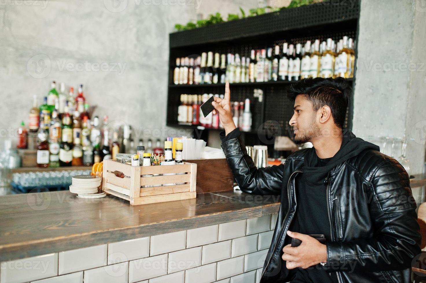 casual en stijlvolle jonge aziatische man draagt een leren jas die op een bar zit en een creditcard vasthoudt om te betalen. foto