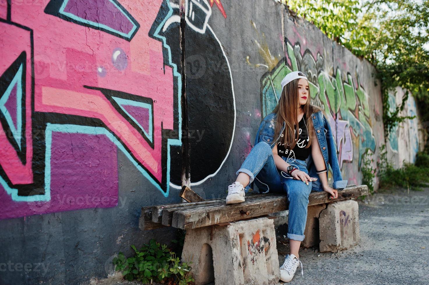 stijlvolle casual hipster meisje in pet en spijkerbroek dragen luisteren muziek van koptelefoon van mobiele telefoon tegen grote graffiti muur met bom. foto