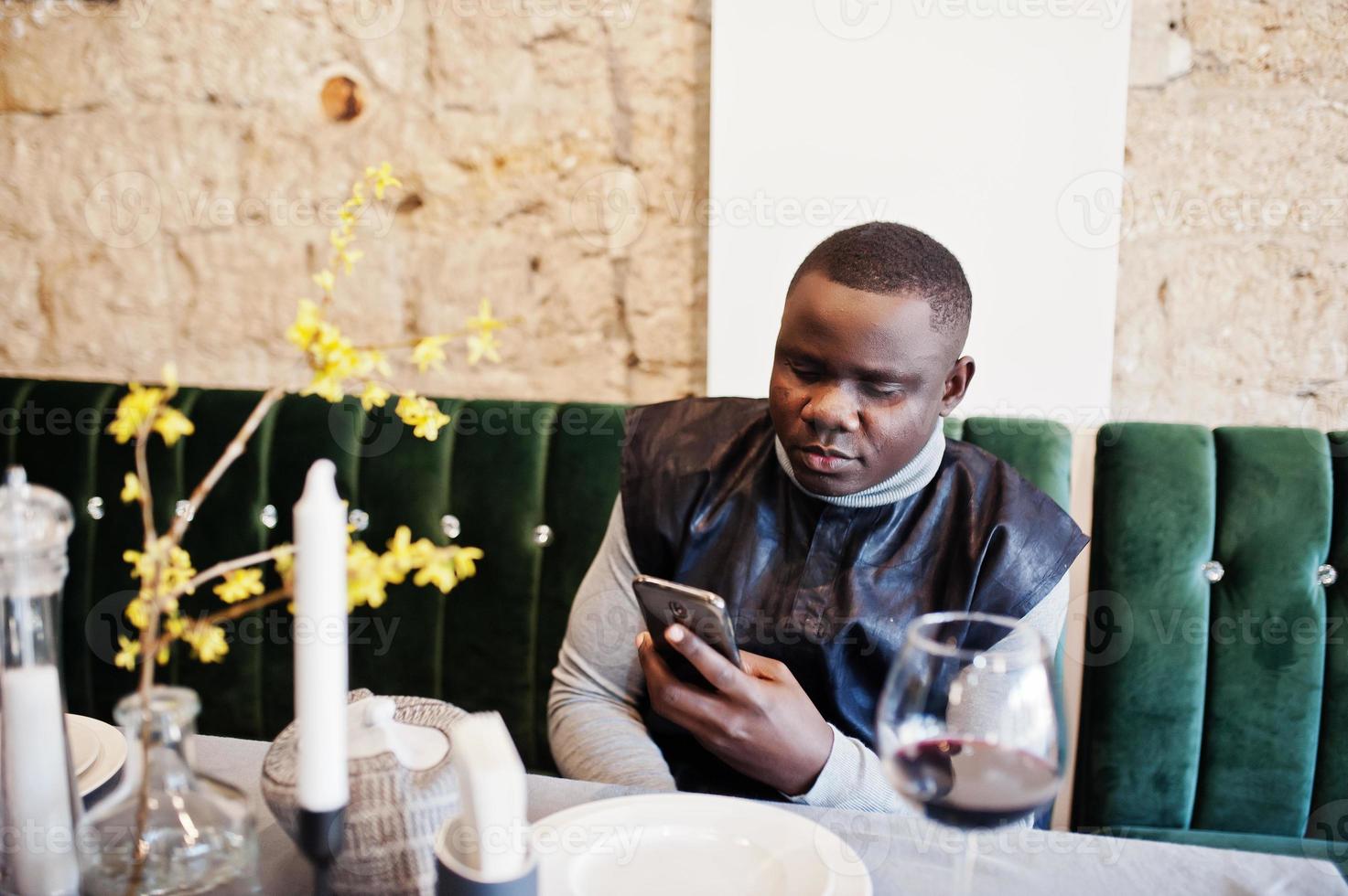 Afrikaanse man in zwarte traditionele kleding zitten in restaurant met glas rode wijn met mobiele telefoon bij de hand. foto