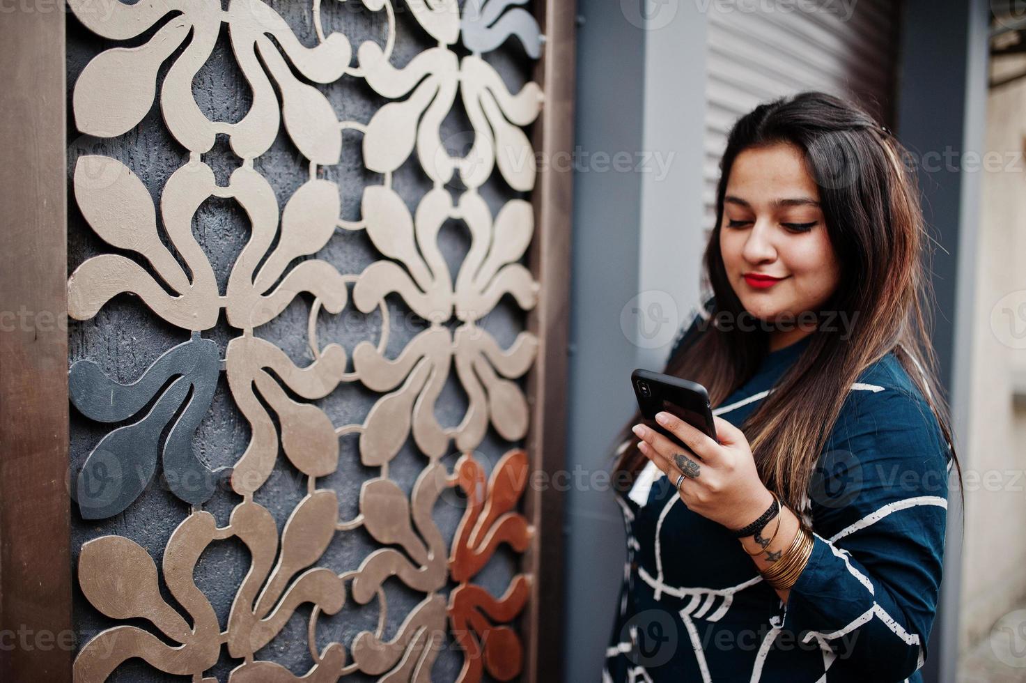 brunette Indiase vrouw in lange modieuze jurk poseerde buiten met mobiele telefoon bij de hand. foto