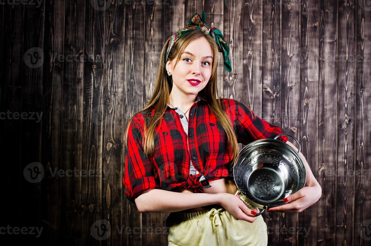 jonge grappige huisvrouw in geruit overhemd en gele korte broek pin-up stijl met keukenzeef op houten achtergrond. foto