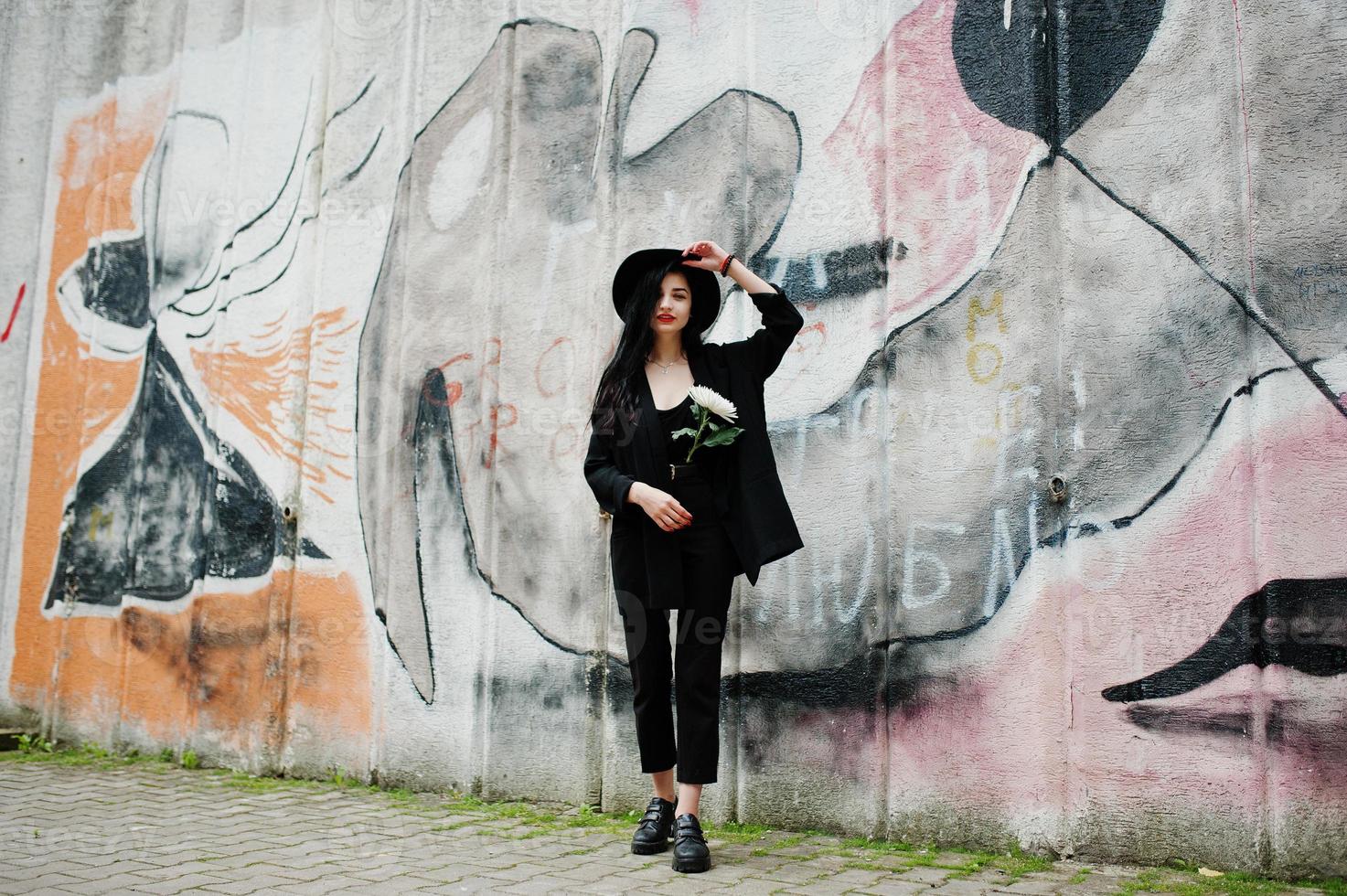 sensueel meisje helemaal in het zwart, rode lippen en hoed. goth dramatische vrouw houdt witte chrysanthemumbloem tegen graffitimuur. foto