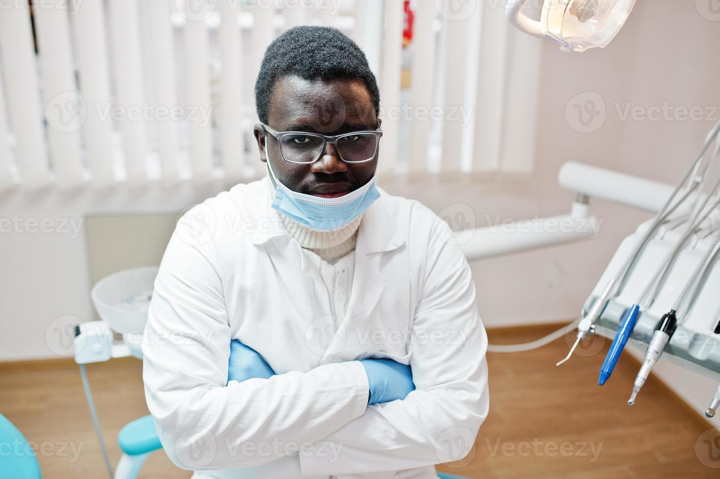 Afro-Amerikaanse mannelijke arts in masker en bril met gekruiste armen zittend op tandartsstoel in tandheelkundige kliniek. foto