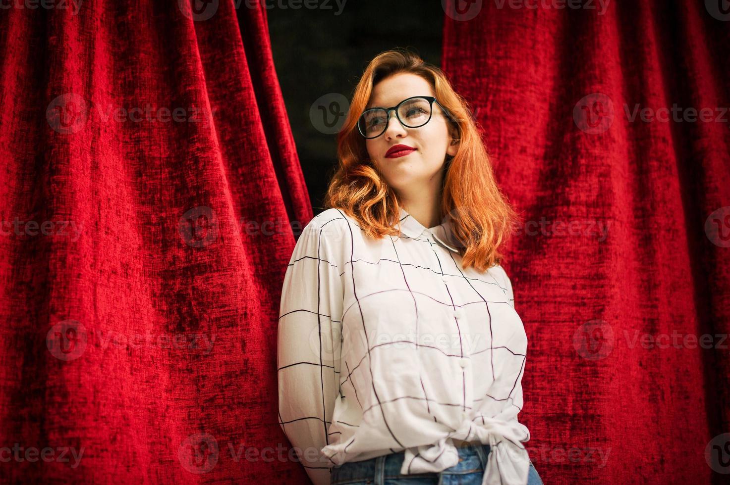 aantrekkelijke roodharige vrouw in brillen, draag op witte blouse poseren bij boog van open rode gordijnen. foto