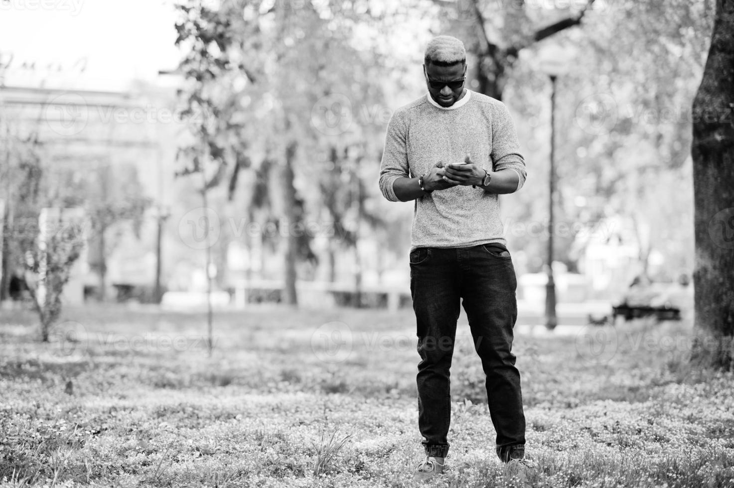 stijlvolle afro-amerikaanse jongen op grijze trui en zwarte zonnebril poseerde in het park en sprak over de telefoon. modieuze zwarte man. foto