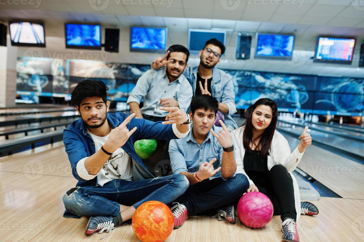 groep van vijf Zuid-Aziatische volkeren die rust en plezier hebben in de bowlingclub, zittend op een bowlingbaan met ballen op handen. foto