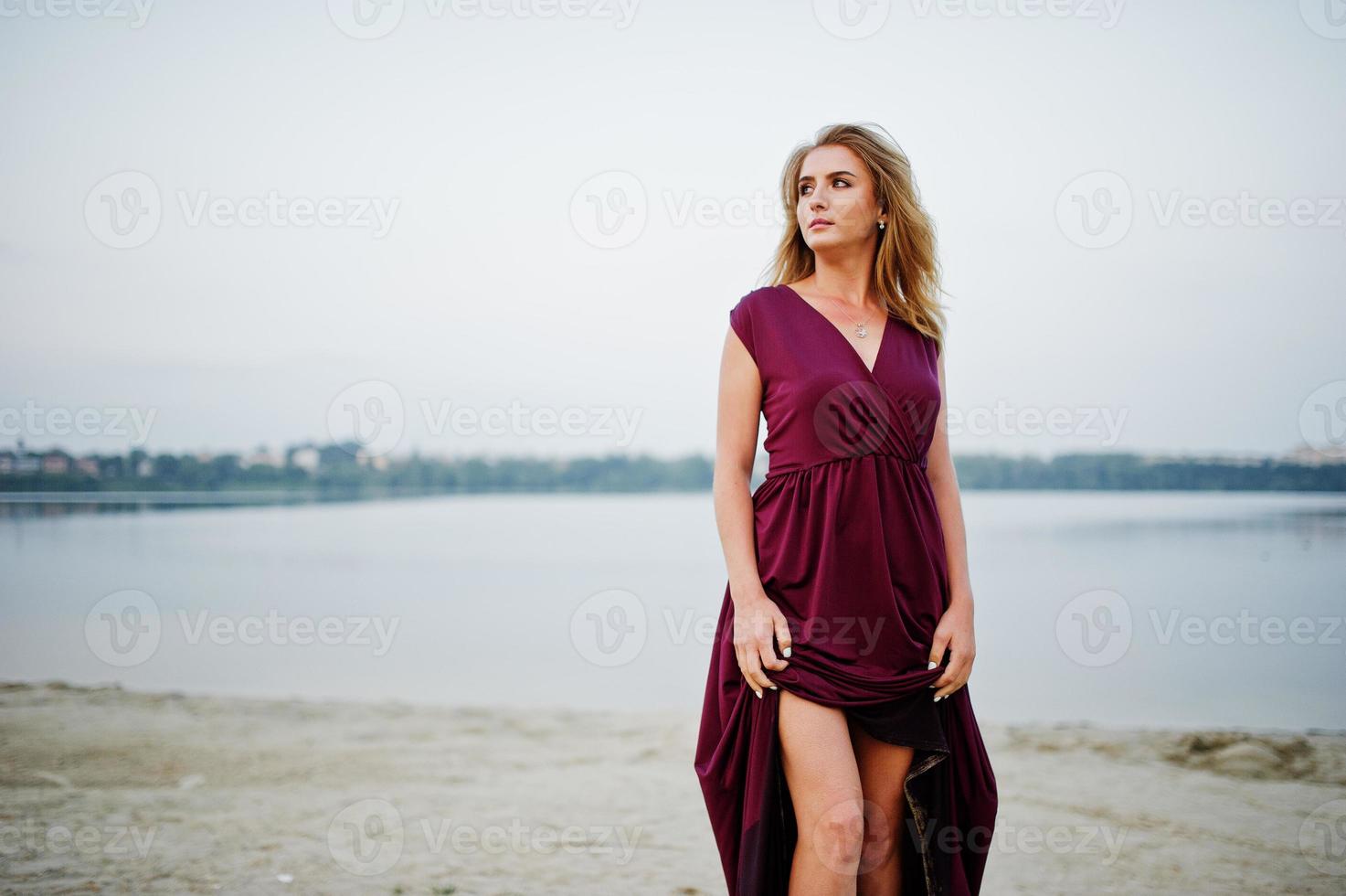 blonde sensuele barefoot vrouw in rode marsala jurk poseren tegen meer op zand. foto