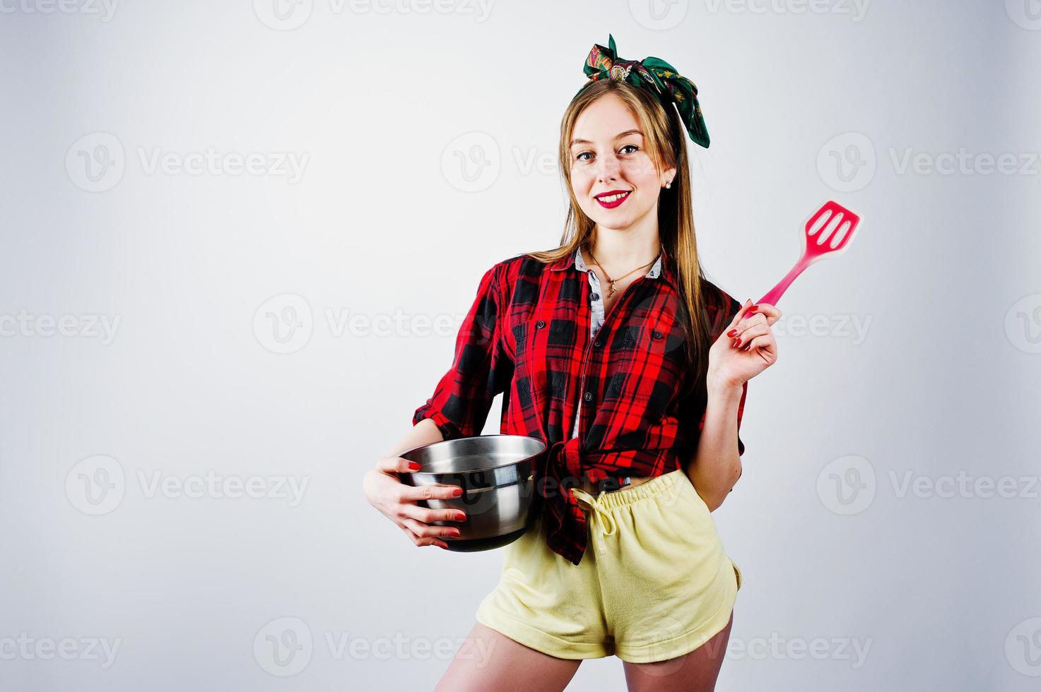 jonge grappige huisvrouw in geruit overhemd en gele korte broek pin-up stijl met pan en keukenlepel geïsoleerd op een witte achtergrond. foto