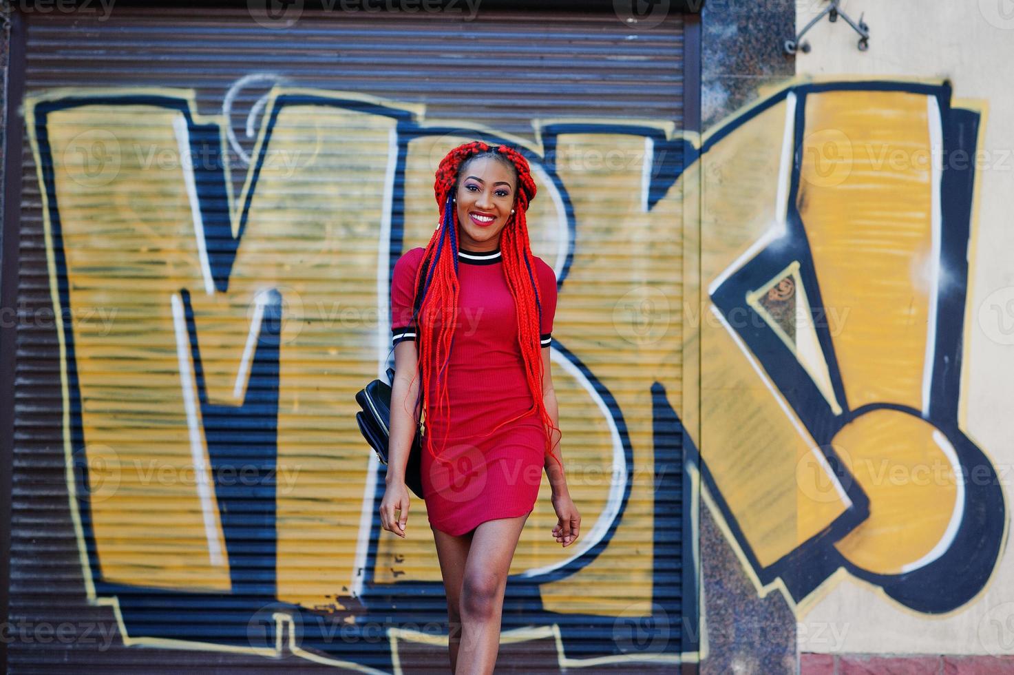 schattig en slank afro-amerikaans meisje in rode jurk met dreadlocks en rugzak poseerde buiten op straat achtergrond graffiti muur. stijlvol zwart model. foto