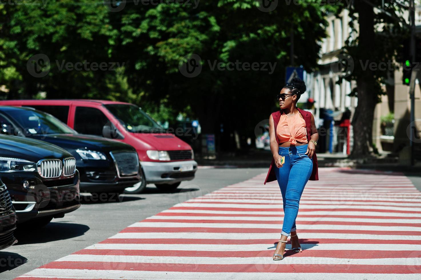 stijlvolle Afro-Amerikaanse vrouw lopen op zebrapad of zebrapad met mobiele telefoon bij de hand. foto