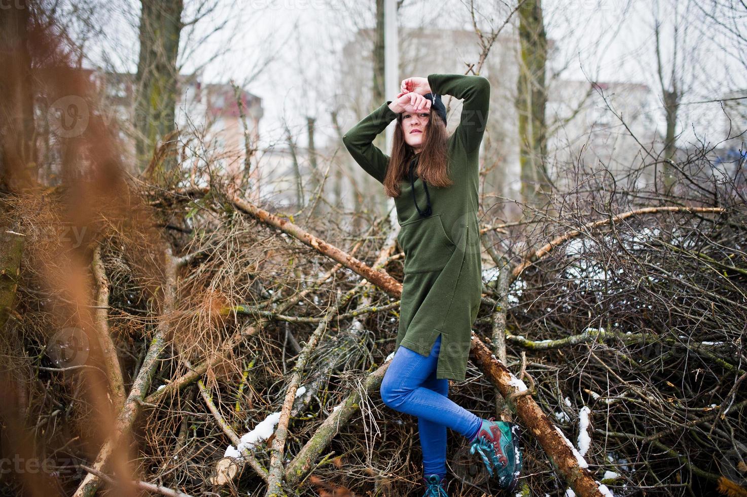 jong meisje draagt een lang groen sweatshirt, jeans en zwarte hoofddeksels aan takken van de dennenboom in de winterdag. foto