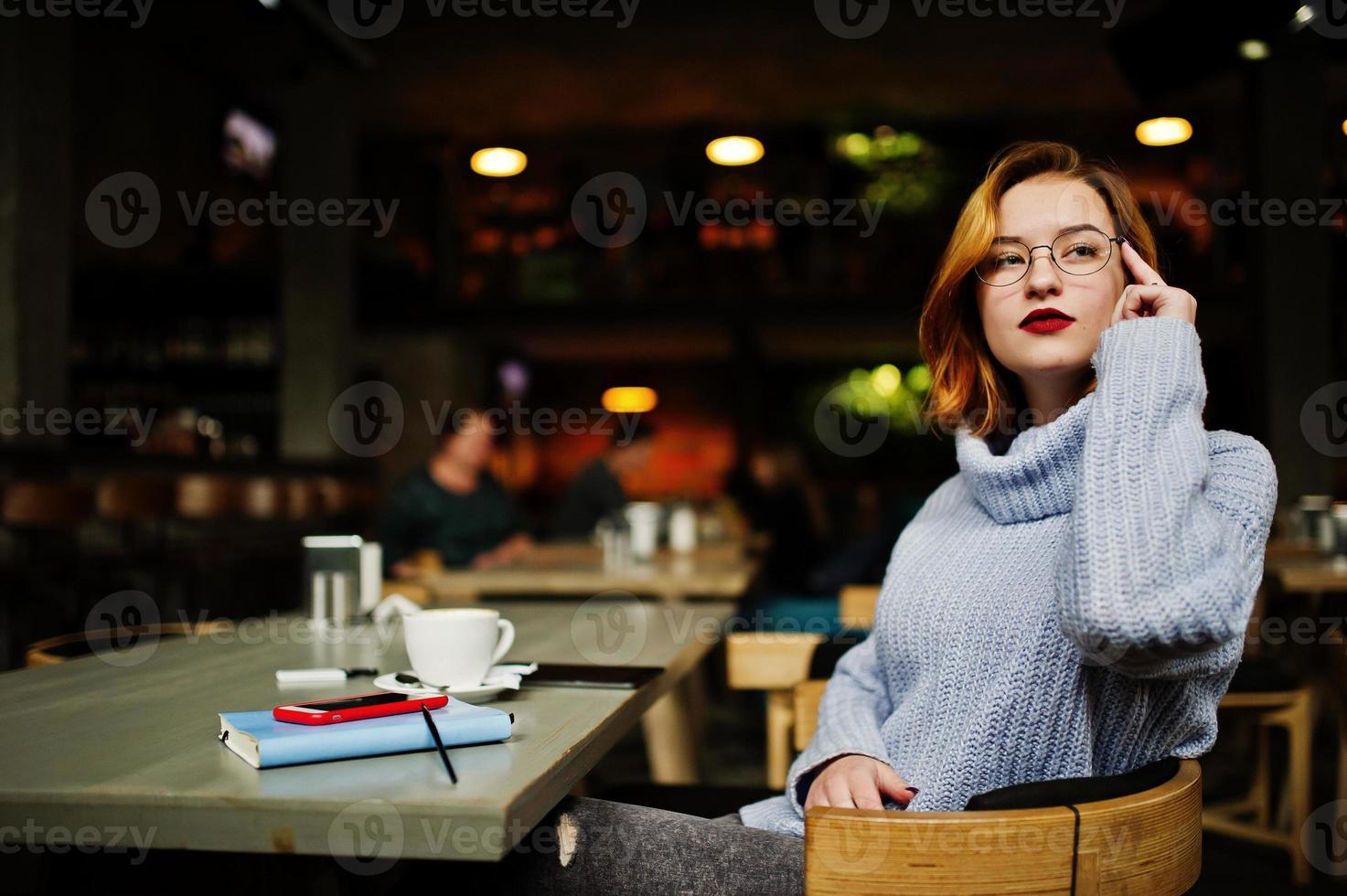 vrolijke jonge mooie roodharige vrouw in glazen zittend op haar werkplek op café met kopje koffie. foto