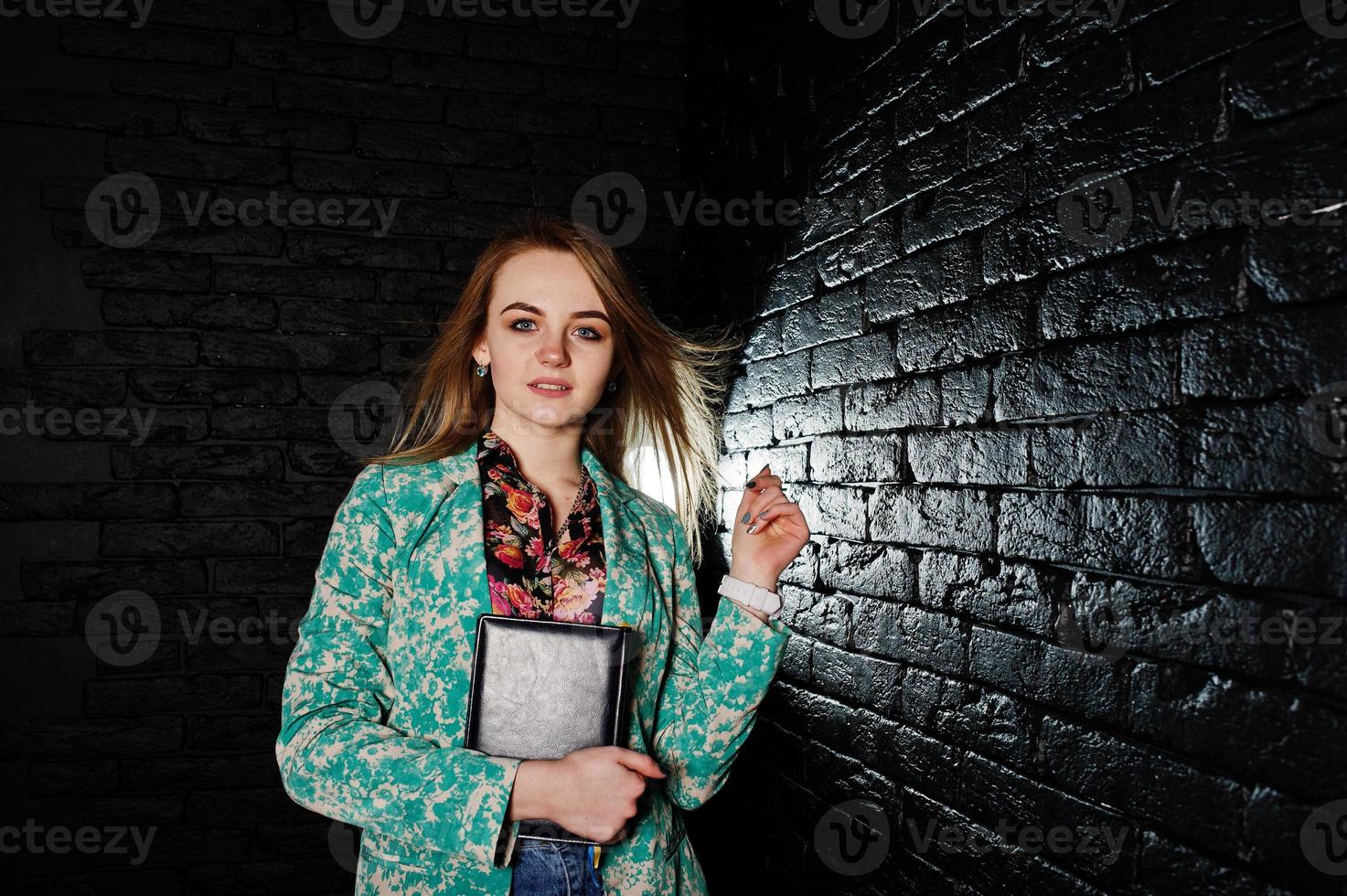 stijlvol blond meisje in jas en spijkerbroek met notebook dagboek bij handen tegen zwarte bakstenen muur in studio. foto