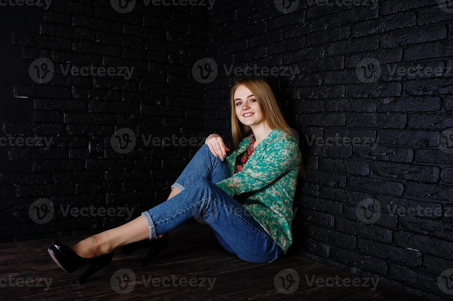 stijlvol blond meisje in jas en spijkerbroek tegen zwarte bakstenen muur in de studio. foto