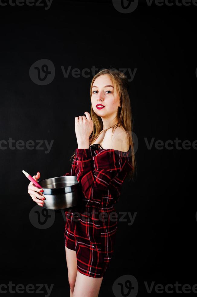 jonge huisvrouw in geruite jurk met pan en keukenlepel geïsoleerd op zwarte achtergrond. foto