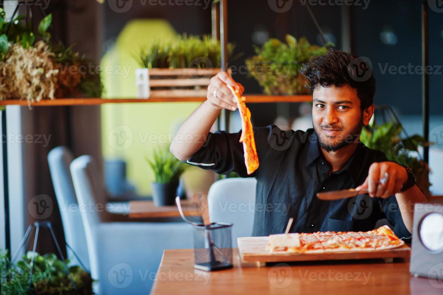 zelfverzekerde jonge indiase man in zwart shirt zit bij pizzeria met pizza. foto