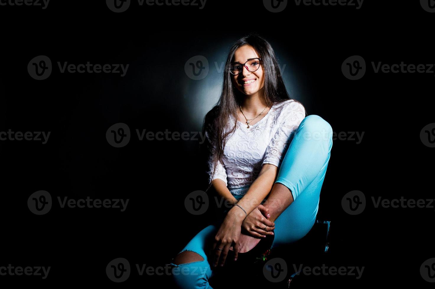 portret van een aantrekkelijke jonge vrouw in witte top en blauwe broek zittend poseren met haar bril in het donker. foto