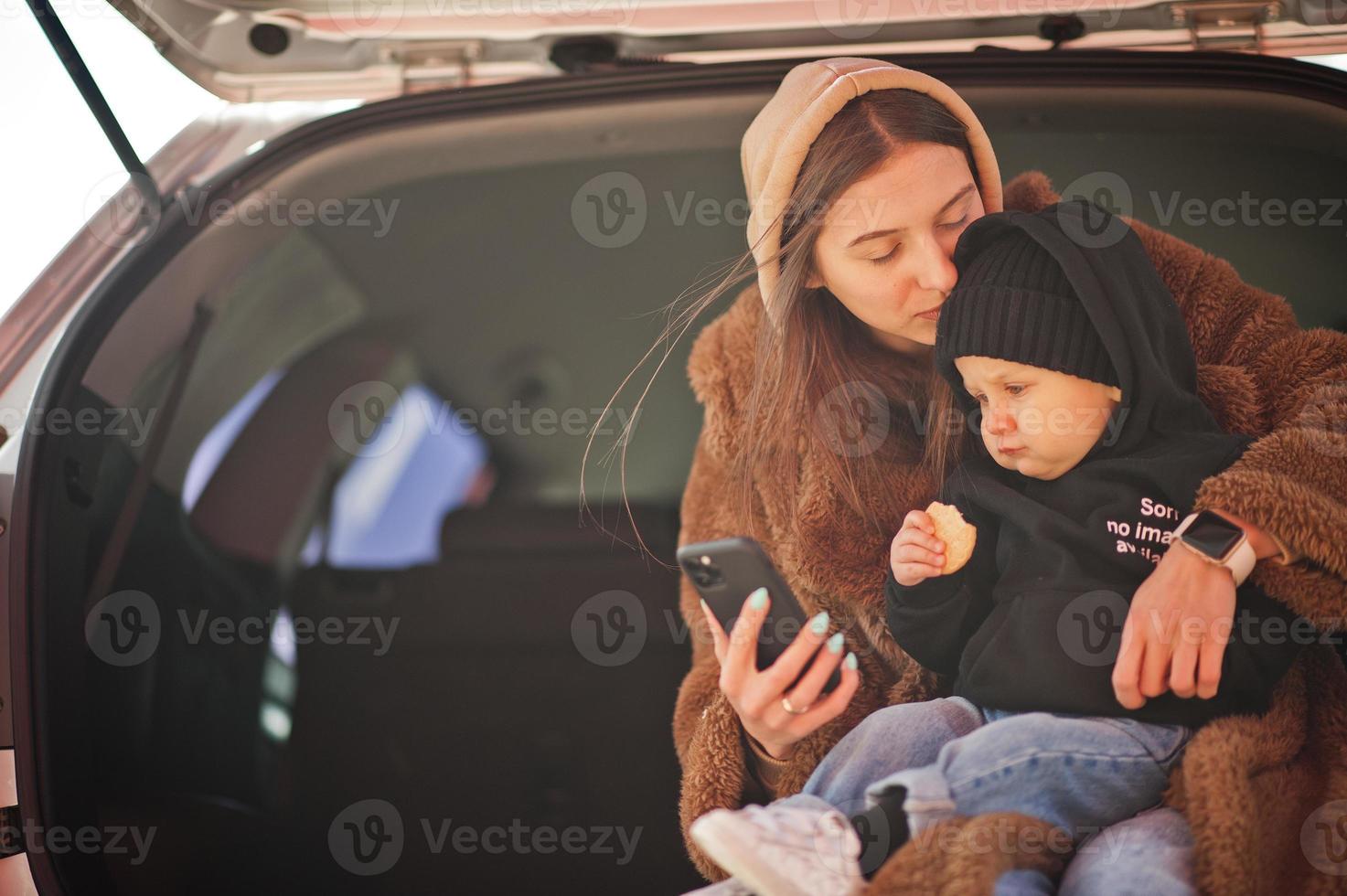 jonge moeder en kind zitten in de kofferbak van een auto en kijken naar mobiele telefoon. veiligheid rijden concept. foto