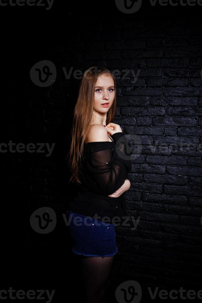 studio shot van brunette meisje in zwarte blouse met beha en korte broek tegen zwarte bakstenen muur. foto