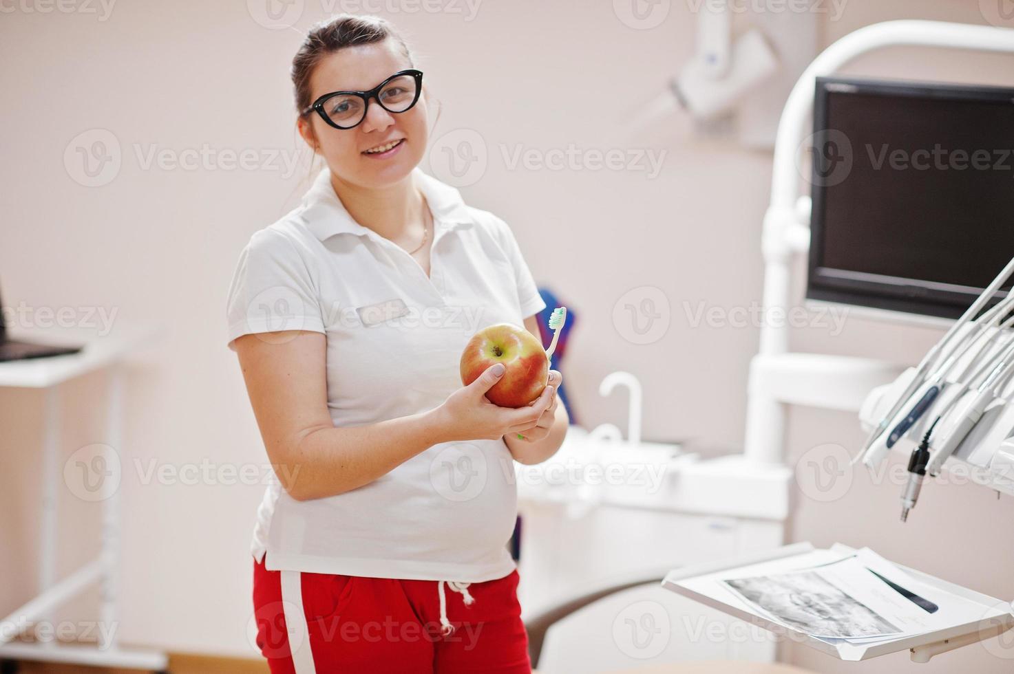 portret van vrouwelijke tandarts vrouw op bril permanent in haar tandheelkunde kantoor met een appel en tandenborstel. foto