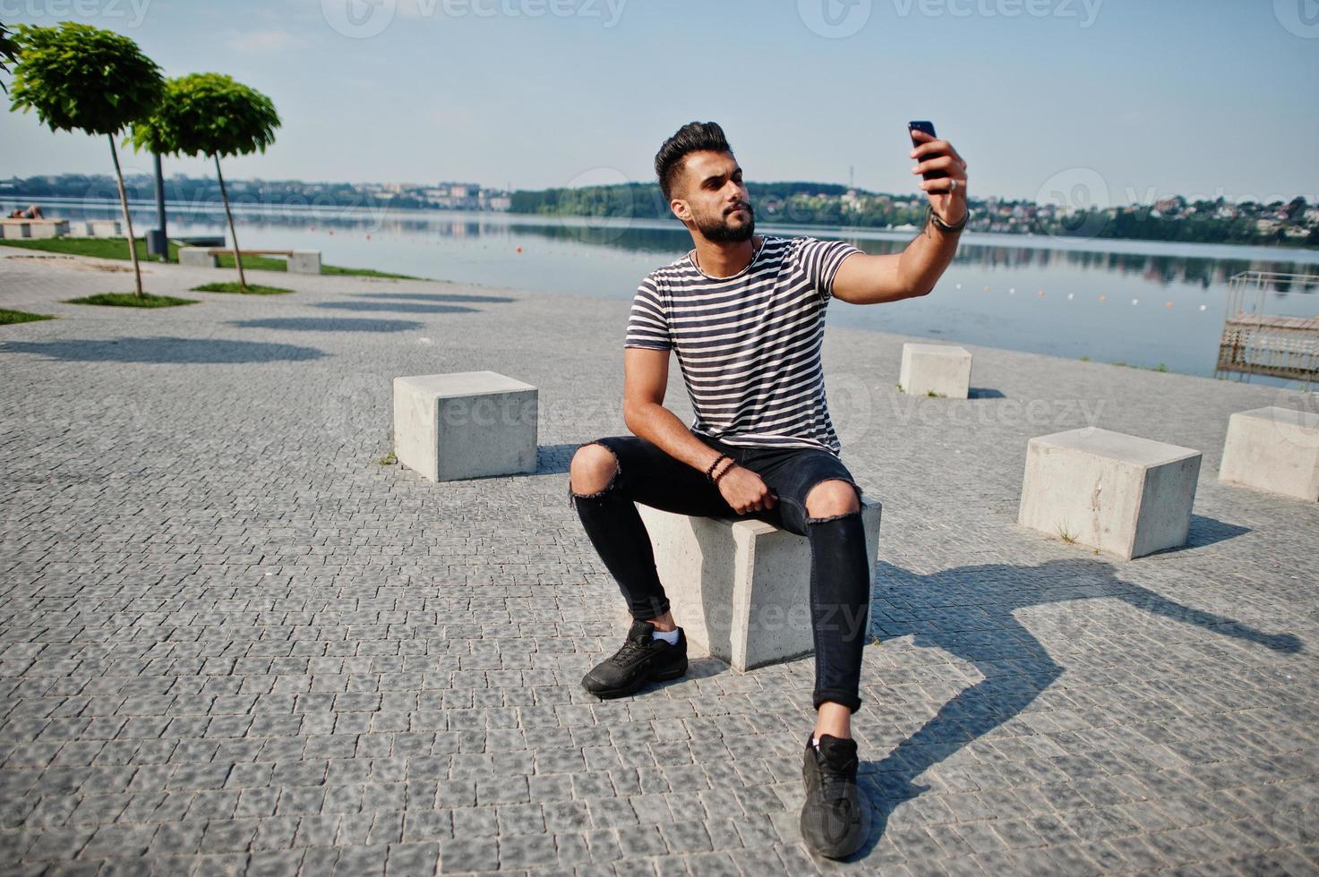 knappe lange Arabische baard man model op gestript shirt buiten geposeerd. modieuze arabische man met mobiele telefoon. foto