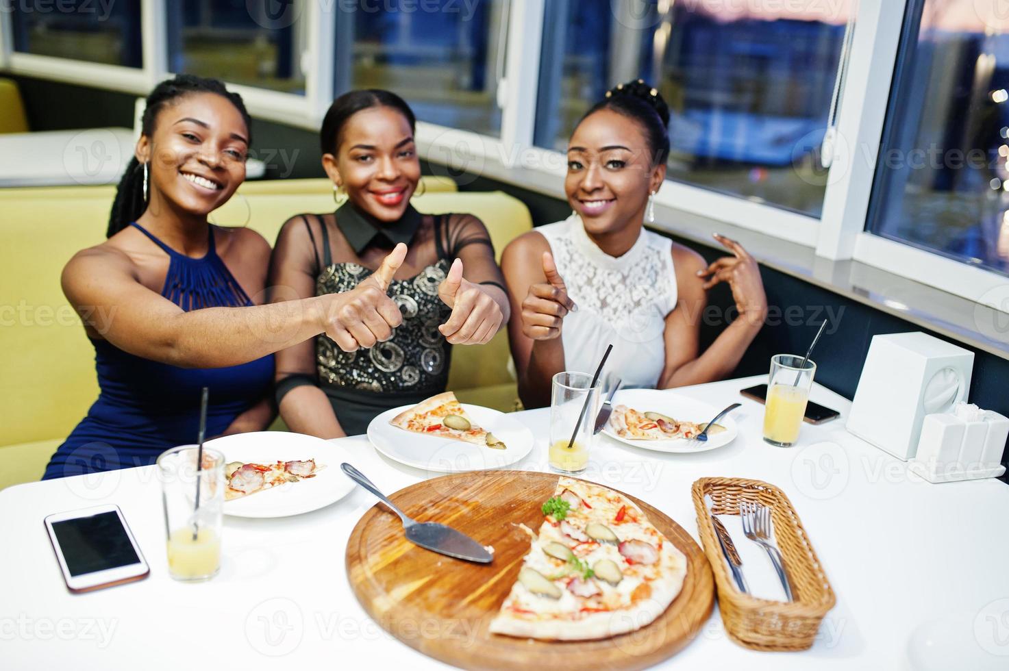drie Afrikaanse vrouw in jurk poseren in restaurant, pizza eten en sap drinken. foto