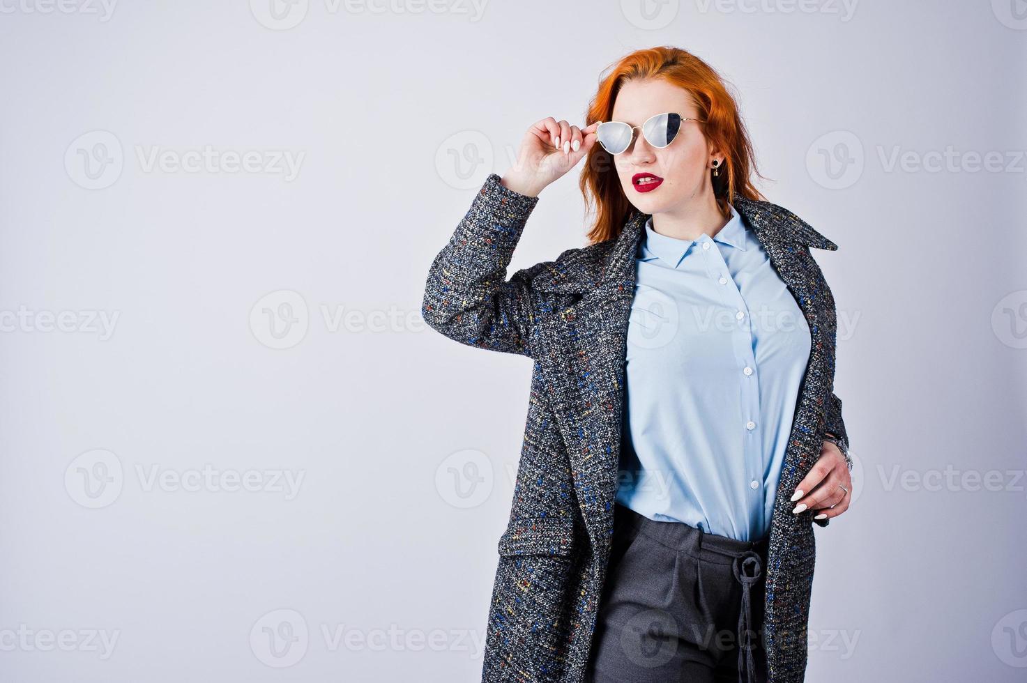 portret van een fantastisch roodharig meisje in blauw shirt, grijze overjas poseren met zonnebril in de studio. foto