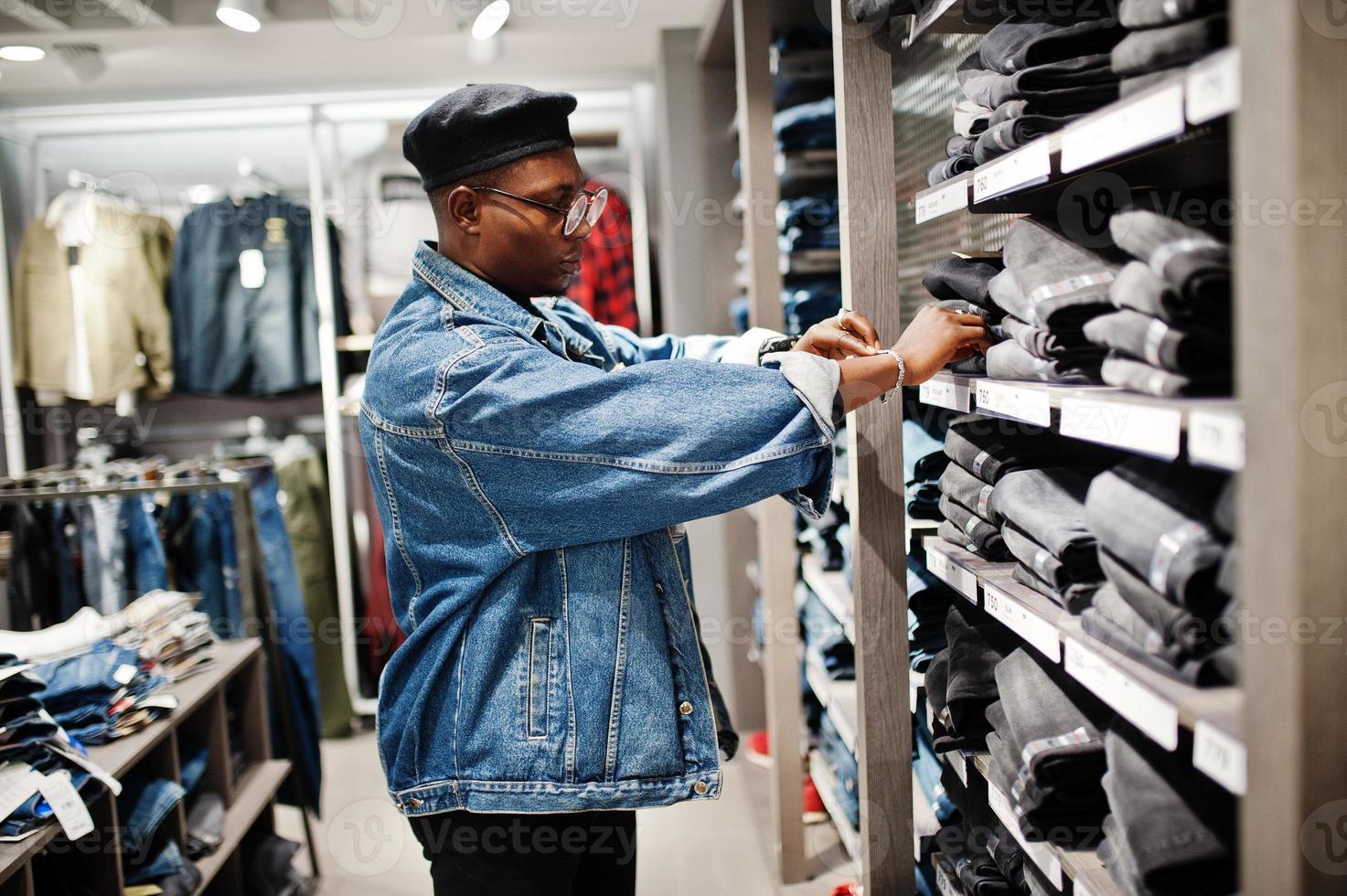 stijlvolle casual Afro-Amerikaanse man bij jeans jasje en zwarte baret bij kledingwinkel kijken op broek op planken. foto