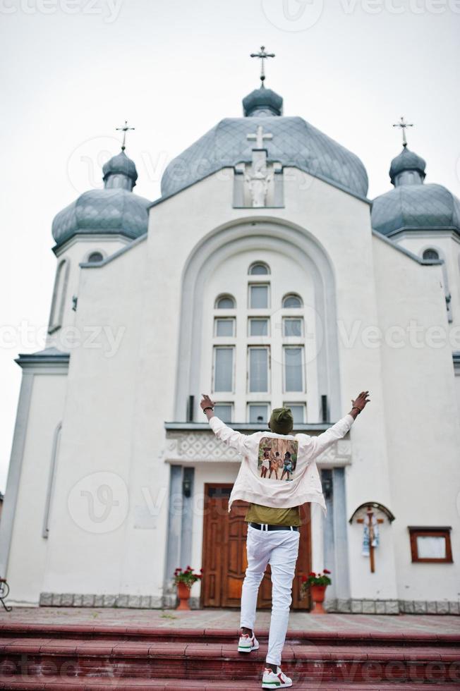 afro-amerikaanse man in hoed staat tegen kerk en steekt handen in de lucht. geloof en christendom in afrika. foto