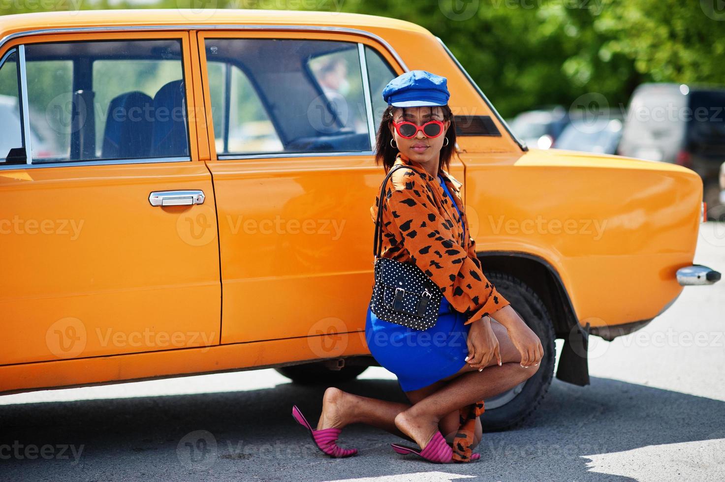 mooie afrikaanse amerikaanse dame met zonnebril die zich dichtbij oranje klassieke retro auto bevindt. foto