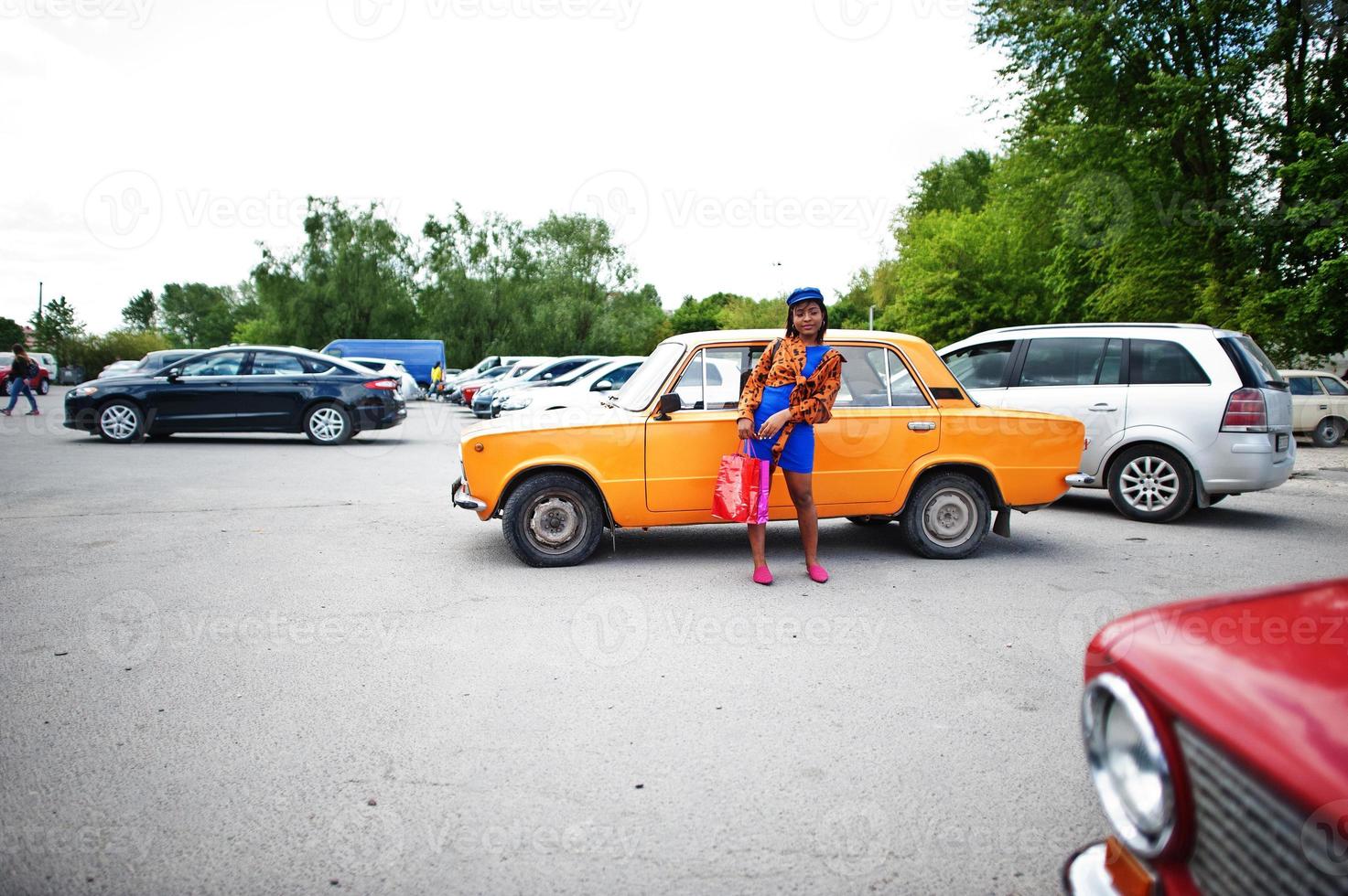 mooie Afro-Amerikaanse dame met boodschappentassen staan in de buurt van oranje klassieke retro auto. foto