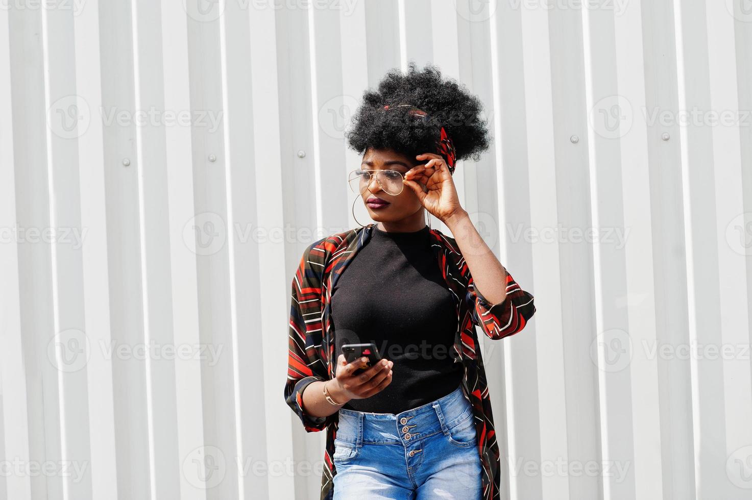 afrikaanse vrouw met afrohaar, in jeansbroek en bril poseerde tegen witte stalen muur met mobiele telefoon in de hand. foto