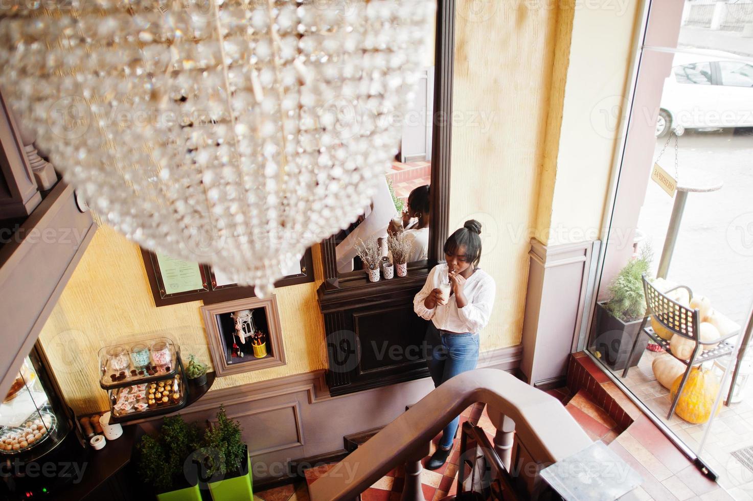 stijlvolle Afro-Amerikaanse vrouwen in witte blouse en spijkerbroek poseerden in café met karamel latte. foto