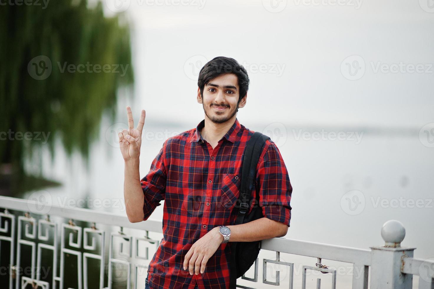 jonge indiase student man op geruit hemd en spijkerbroek met rugzak gesteld op avond stad tegen meer, toont twee vingers. foto