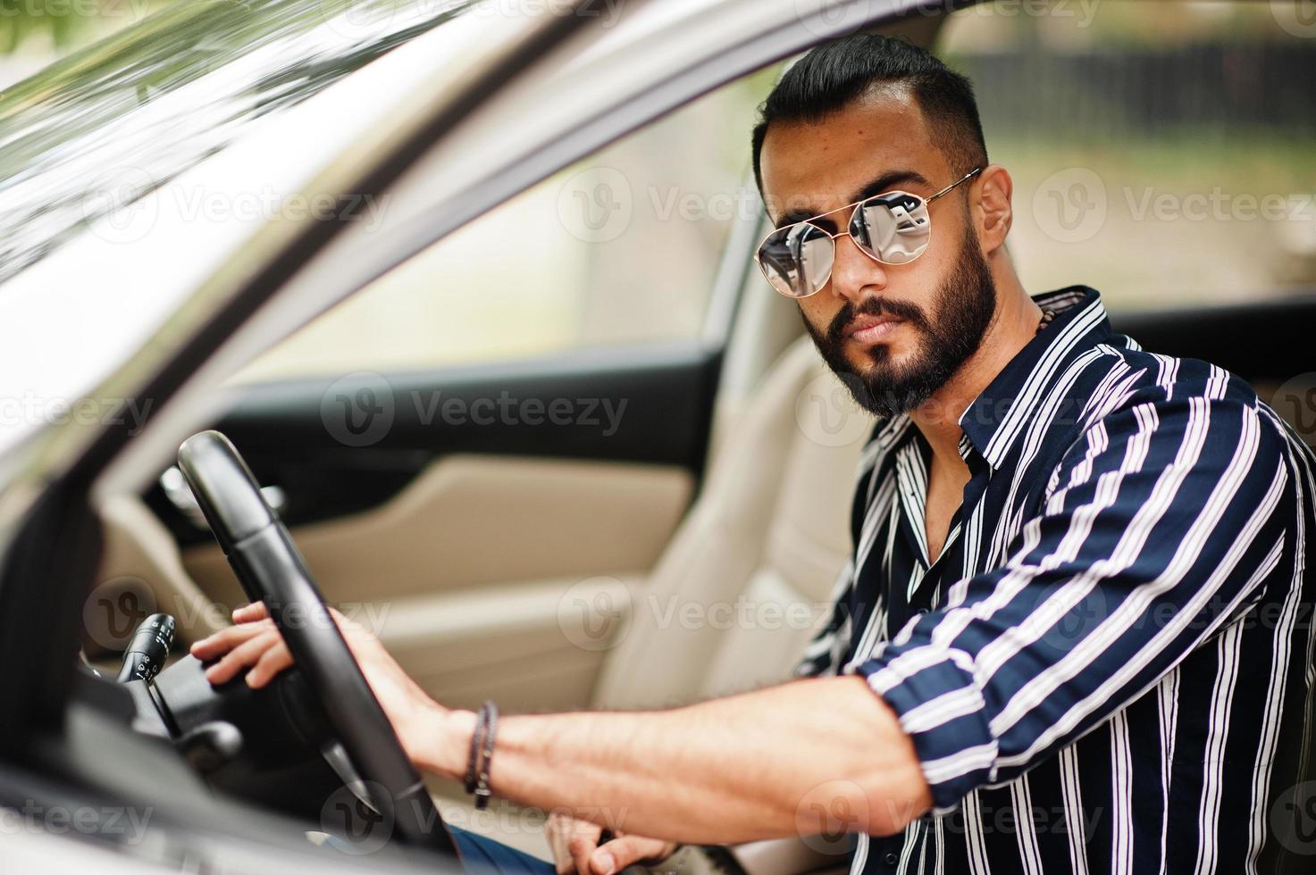 succesvolle arabische man draagt een gestreept shirt en een zonnebril poseert achter het stuur van zijn witte suv-auto. stijlvolle arabische mannen in transport. foto