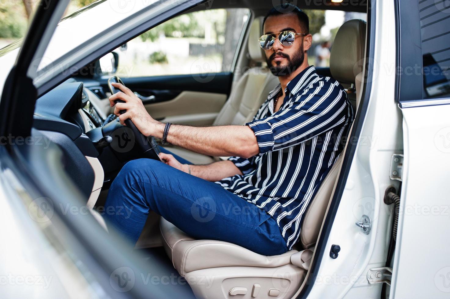 succesvolle arabische man draagt een gestreept shirt en een zonnebril poseert achter het stuur van zijn witte suv-auto. stijlvolle arabische mannen in transport. foto