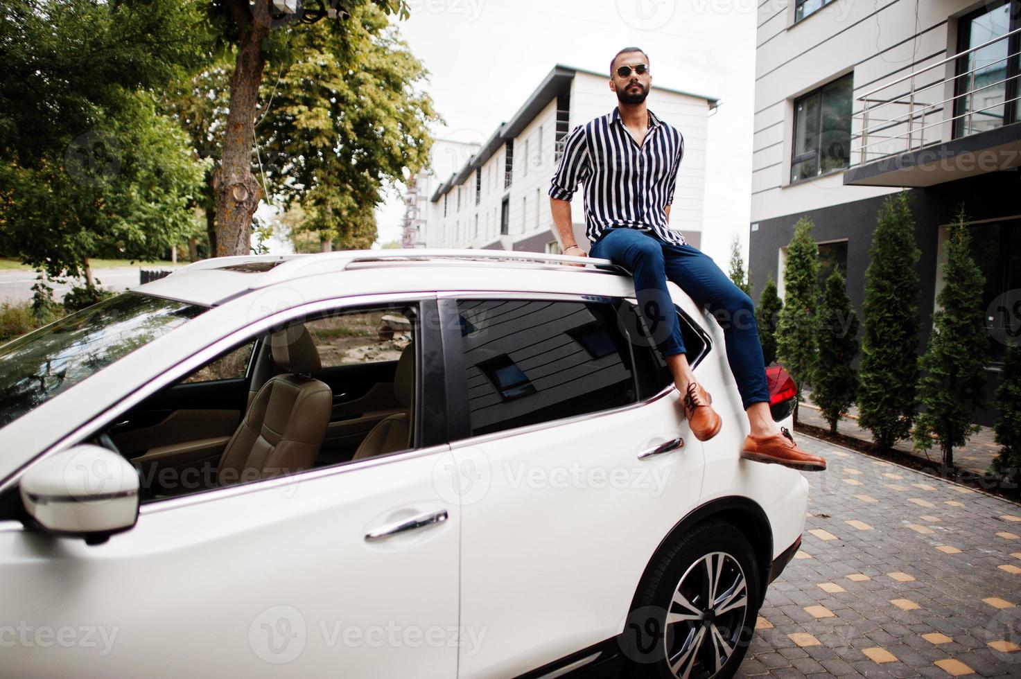 succesvolle arabische man draagt een gestreept shirt en een zonnebril die op het dak van zijn witte suv-auto zit. foto