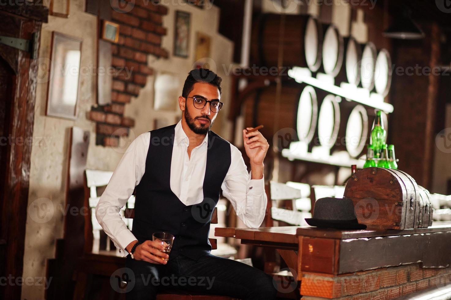 knappe goed geklede arabische man met glas whisky en sigaar poseerde in de pub. foto