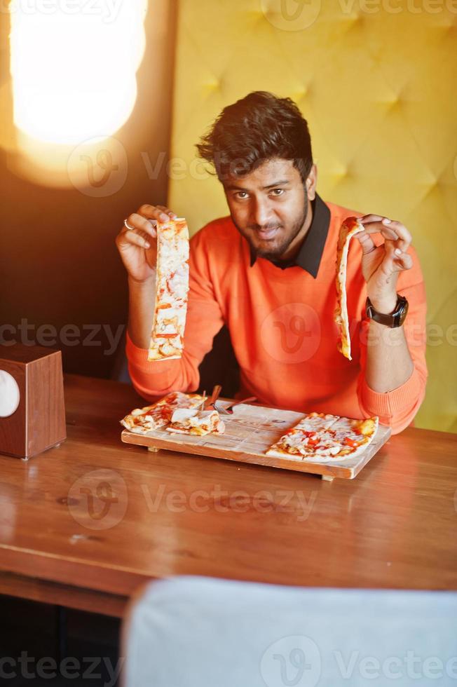 zelfverzekerde jonge indiase man in oranje trui zit bij pizzeria en eet pizza. foto