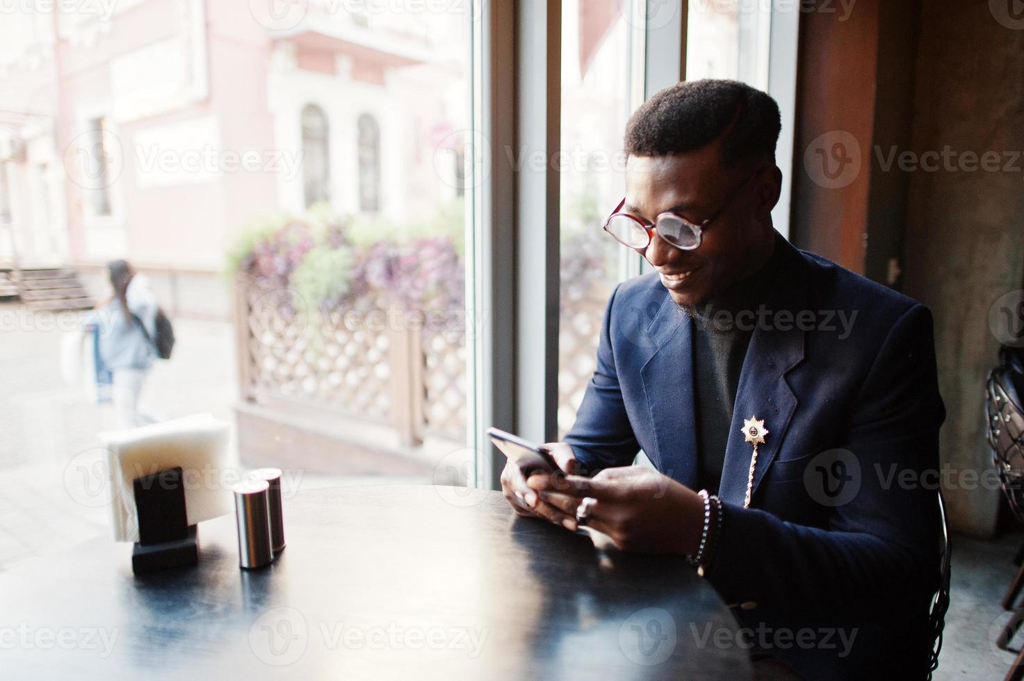 modieuze Afro-Amerikaanse man in pak en bril met mobiele telefoon in de hand op café. foto