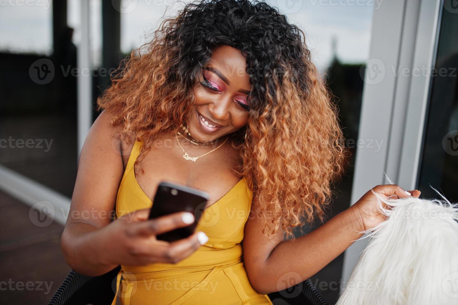 glamour Afro-Amerikaanse vrouw in gele jurk en witte wollen cape zittend op een stoel en kijken op mobiele telefoon. foto