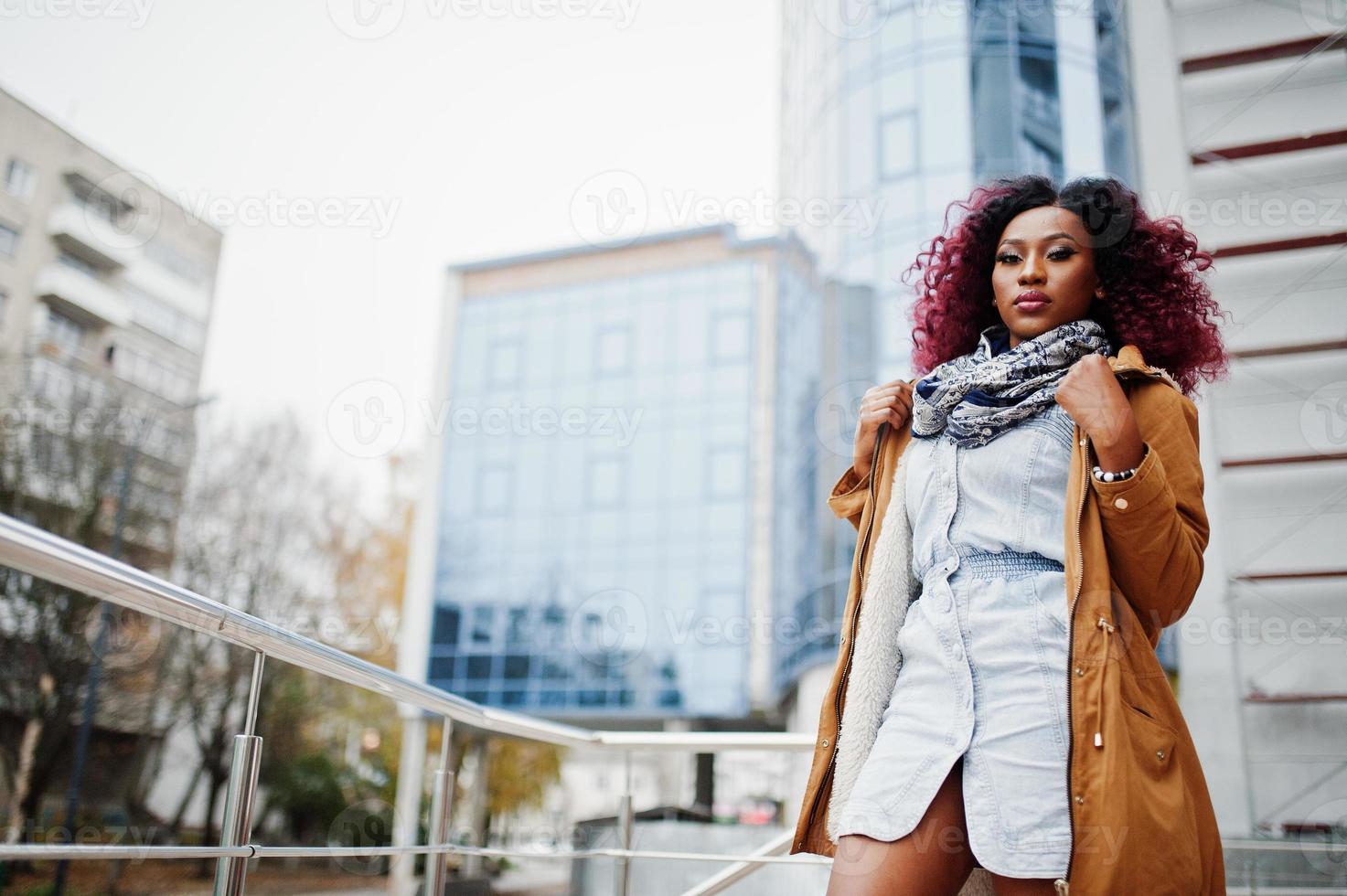 aantrekkelijke krullende Afro-Amerikaanse vrouw in bruine jas poseerde in de buurt van balustrades tegen modern gebouw met meerdere verdiepingen. foto